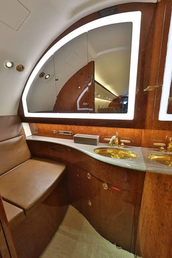 An aircraft lavatory features a gold sink, polished wood paneling, and a large mirror with accent lighting.