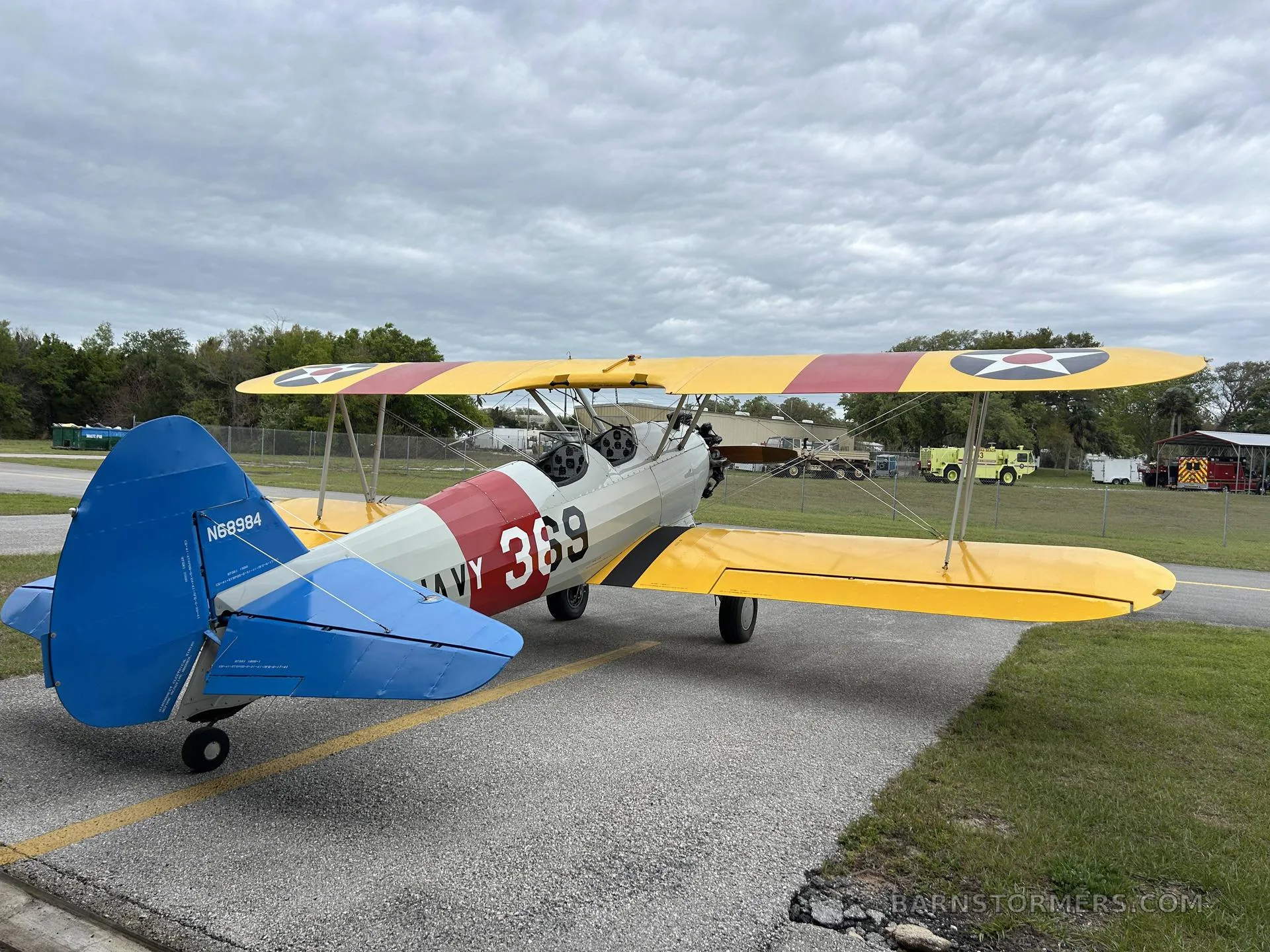 A yellow and white biplane with 'US NAVY 369' markings and a blue tail, seen from the rear-left on an asphalt ramp.