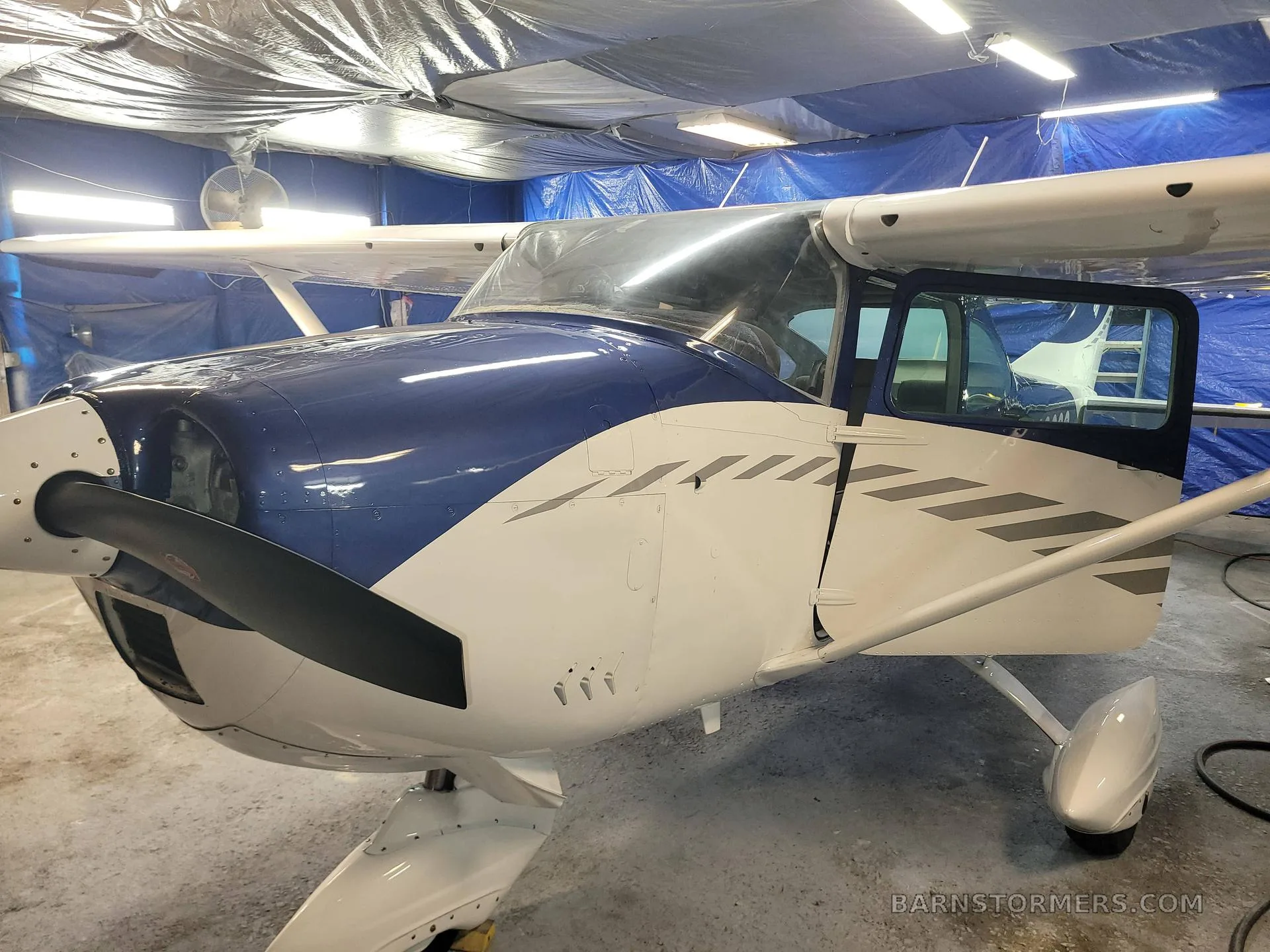 A blue and white aircraft with an open door, showing the front fuselage and propeller in a hangar.