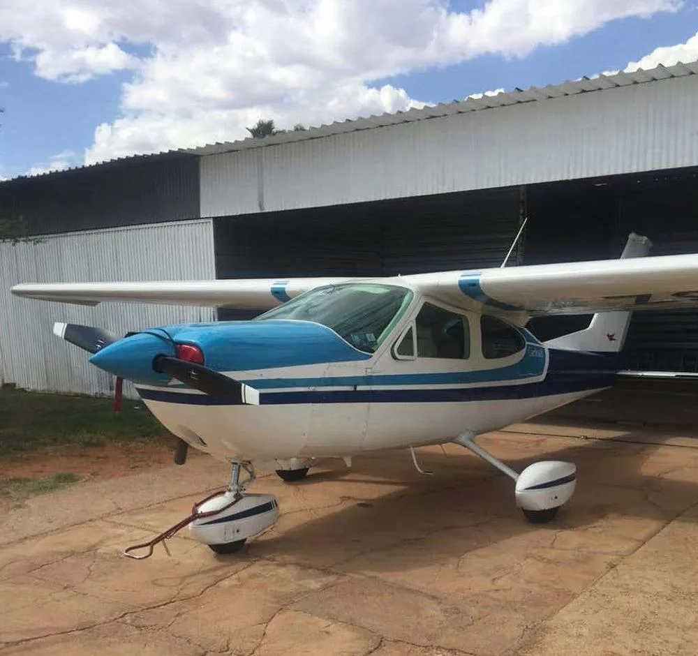 A white and blue Cessna 210 Centurion aircraft parked on a dirt ground in front of an open hangar.
