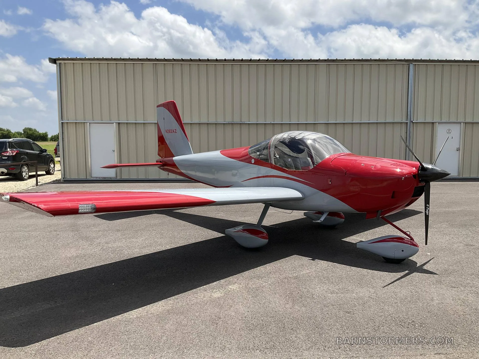 A red and grey single-engine aircraft with a propeller, parked on an asphalt surface in front of a beige hangar.