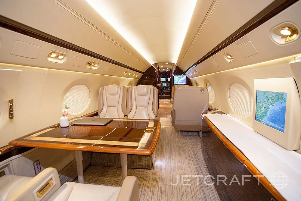A wide view of the private jet's cabin, showing a large wooden table, beige seats, and a screen displaying a map.