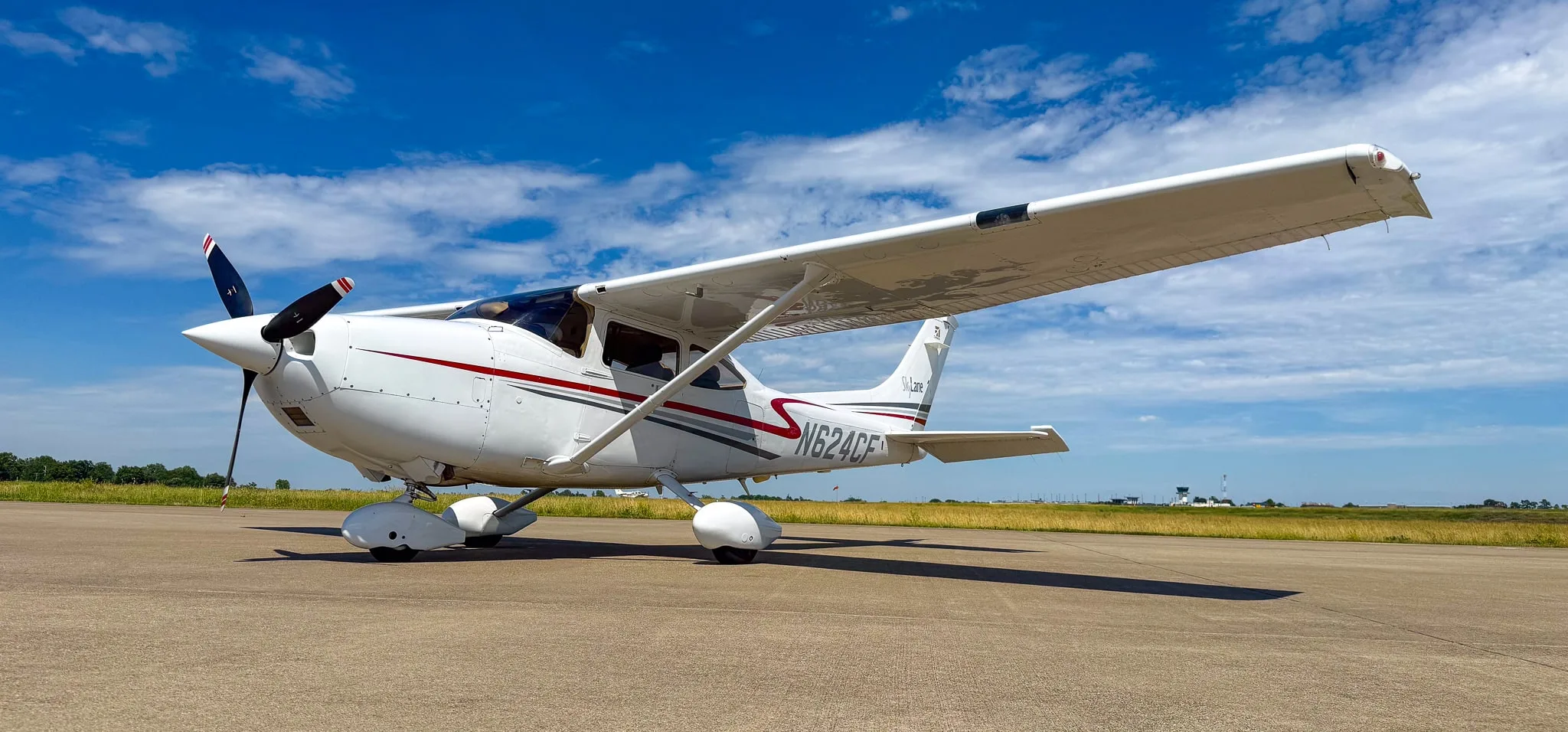 A white Cessna 182 Skylane with red and gray stripes, registration N624CF, parked on an asphalt ramp under a blue sky.
