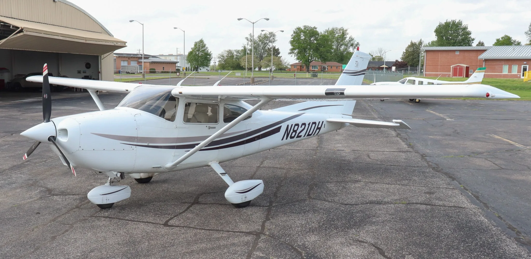 A white Cessna Skylane N821DH is parked on an asphalt tarmac next to a hangar, with another small aircraft in the background.