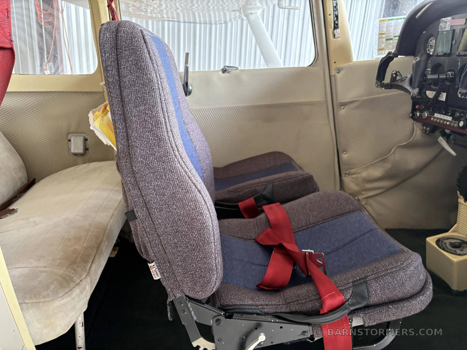 The interior of a small aircraft cabin, showing two front seats with blue and gray fabric and red seatbelts.