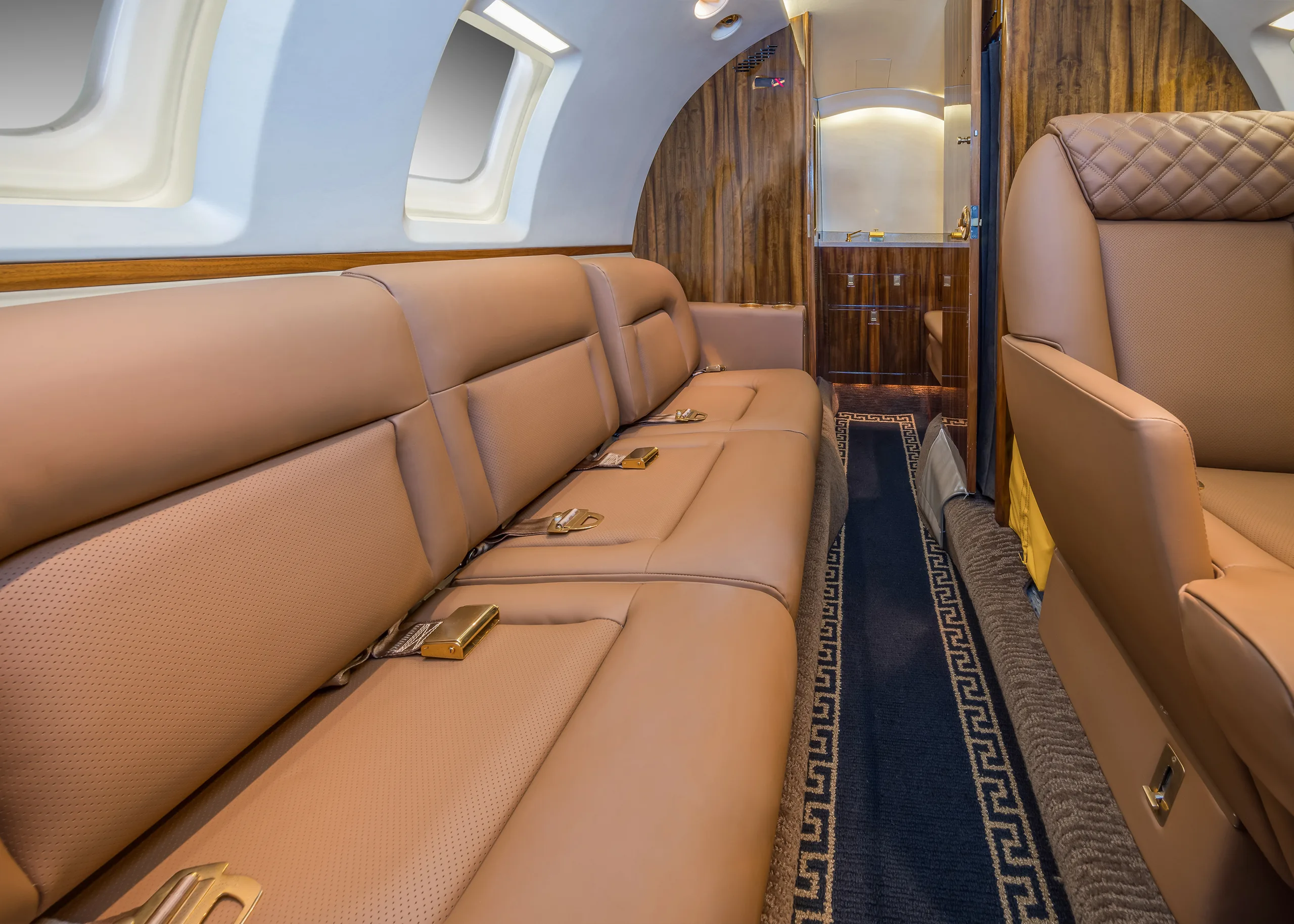 A close-up view of the tan leather couch seating inside a private jet cabin, with a galley area visible in the background.