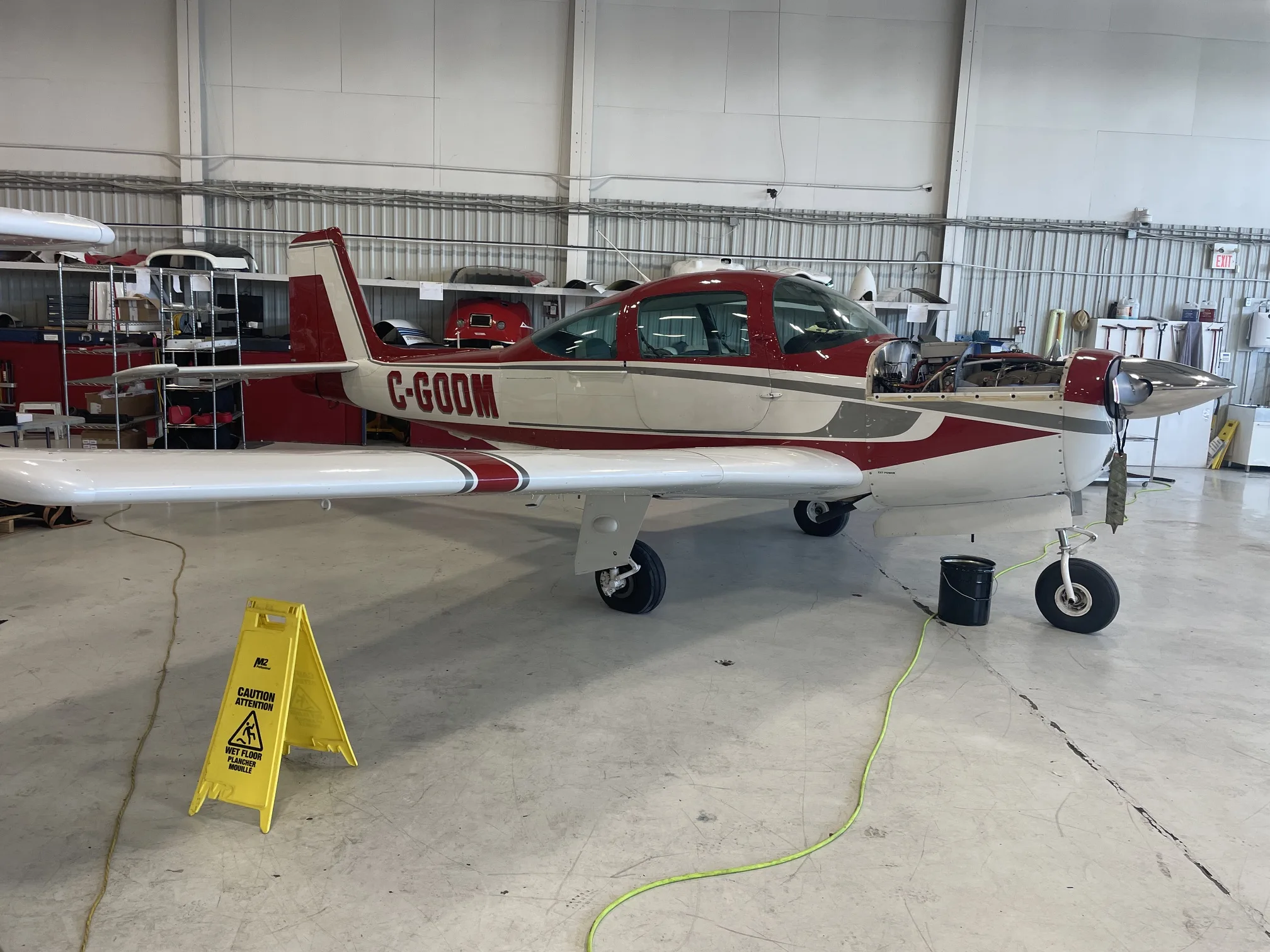 A red and white single-engine aircraft, registration C-GODM, with its engine cowling open, parked in a hangar.