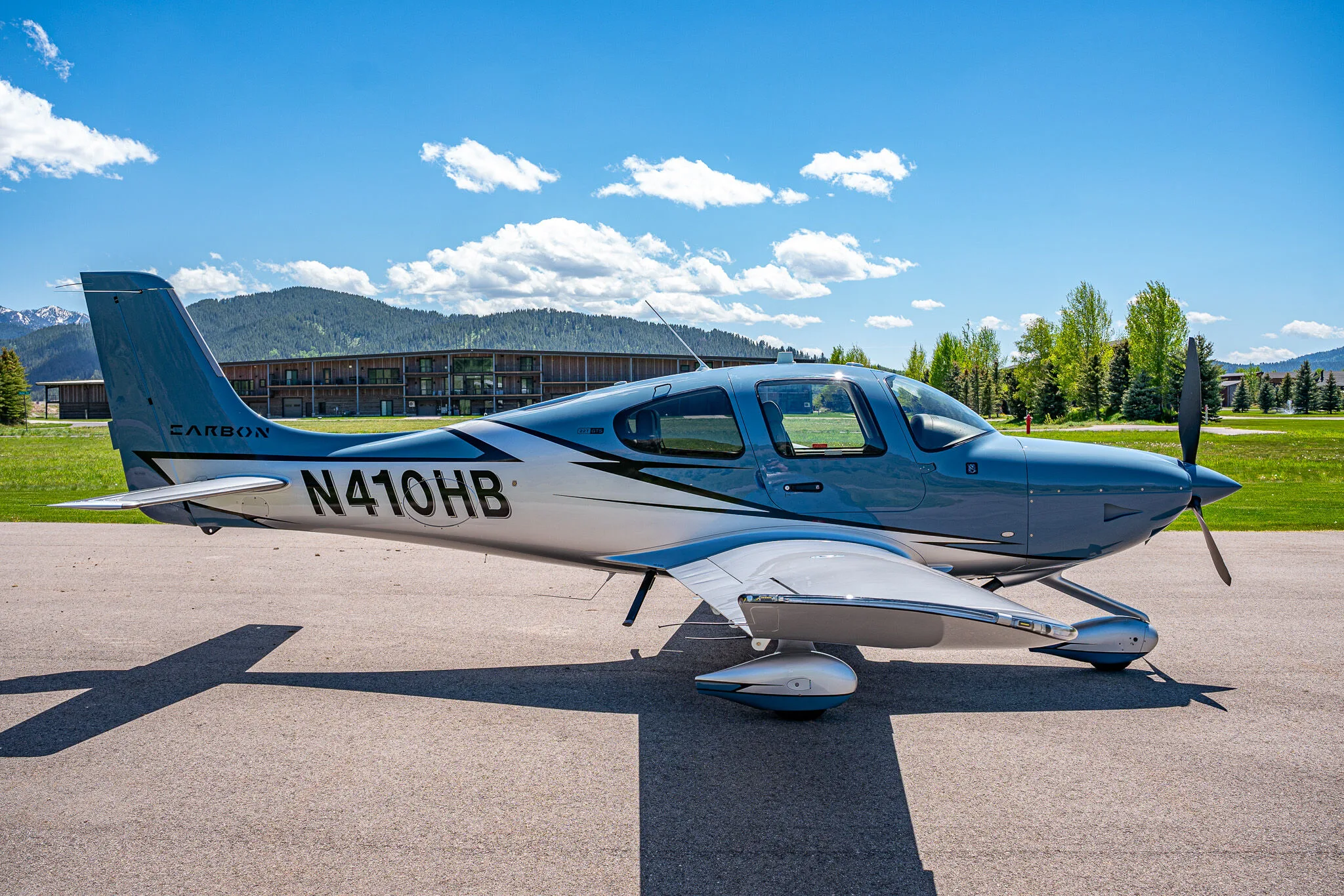 A light blue and silver Cirrus SR22T aircraft (N410HB) with 'CARBON' livery parked on an airfield with buildings and trees in the background.
