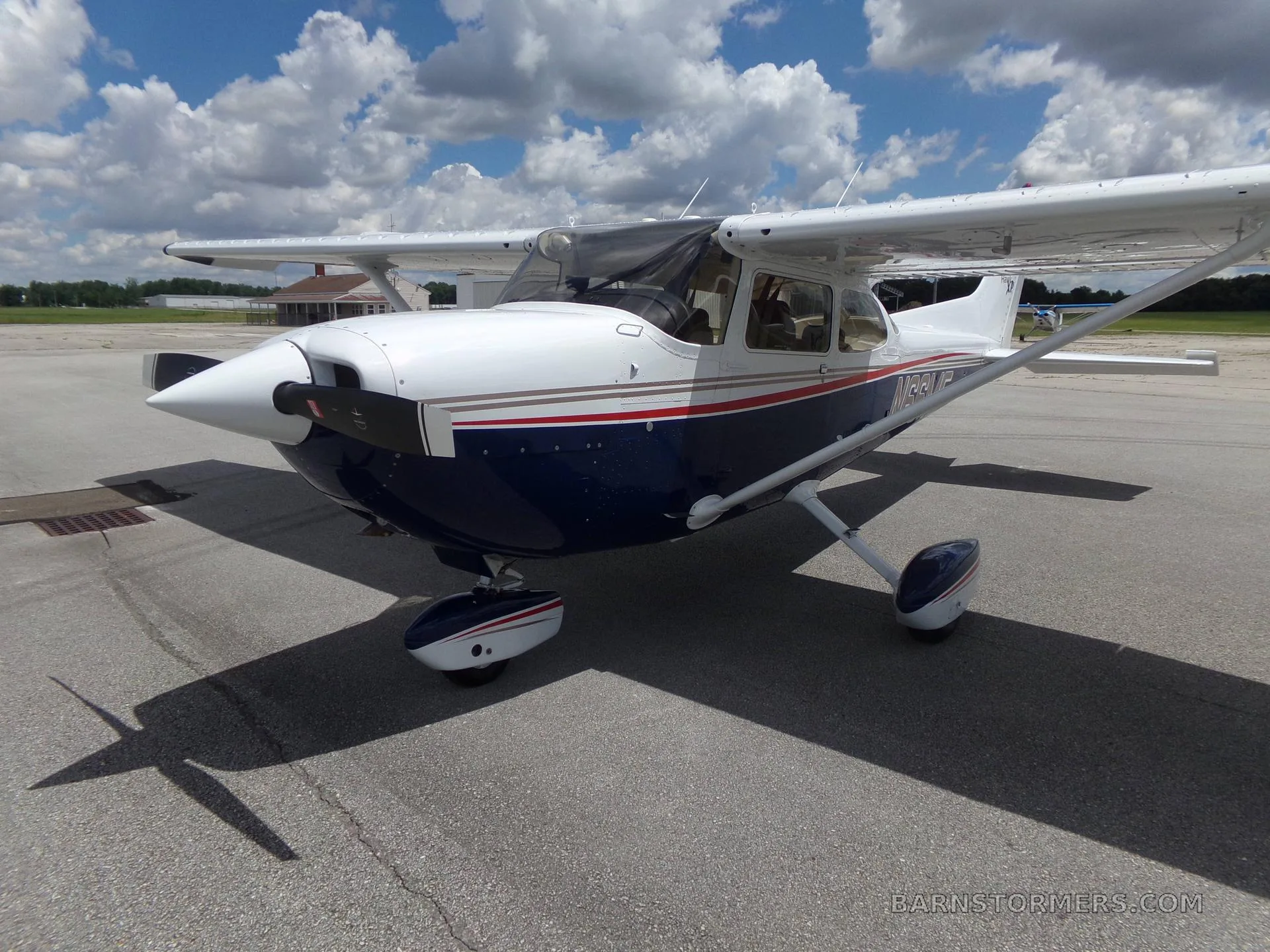 A white and blue Cessna 172 Hawk XP with red and gold stripes, seen from the front-right on an airfield under a partly cloudy sky.