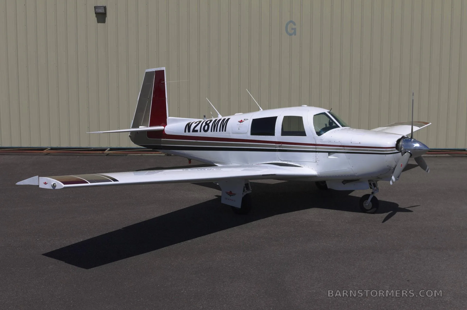 A white single-engine propeller aircraft with red and brown stripes on the tail and wings, parked on an asphalt surface in front of a light-colored hangar.