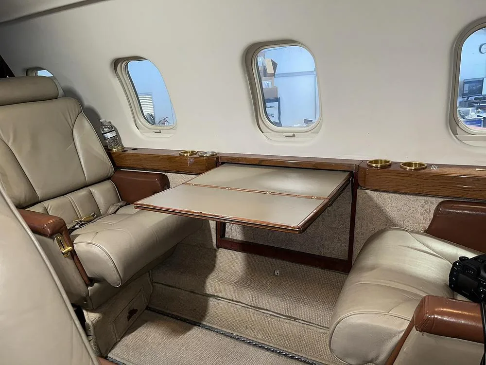 The interior cabin of a private jet with beige leather seats, a fold-out table, and windows.