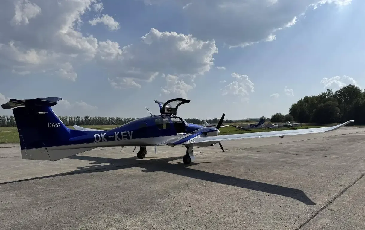 A rear-side view of a blue and silver Diamond DA62 aircraft with registration OK-KEV and open doors on a tarmac.