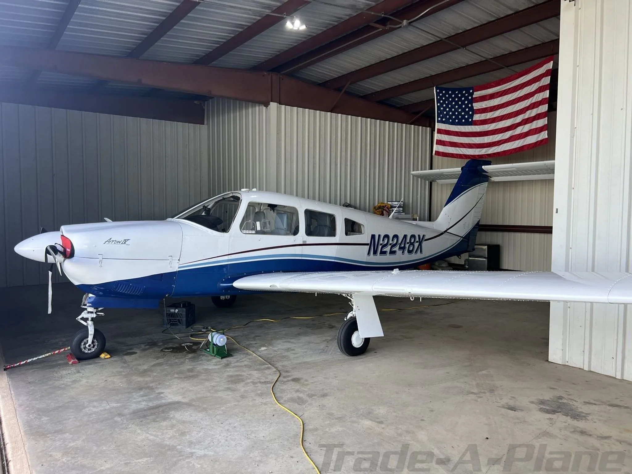 A white and blue Piper Archer II (N2248X) parked inside a hangar with an American flag hanging.