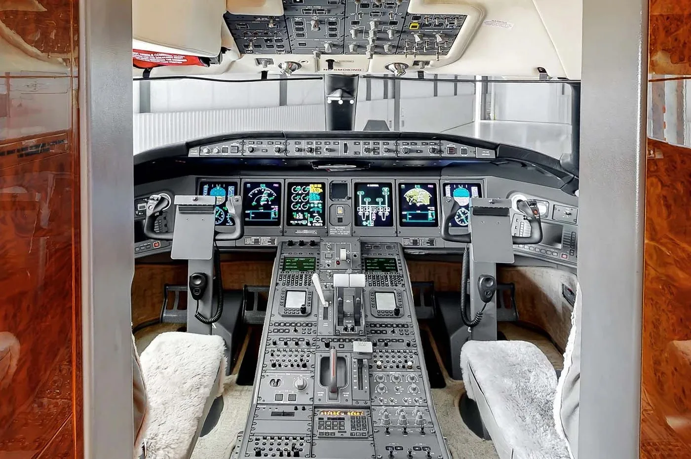 The cockpit of a business jet features multiple digital displays, a complex control panel, and two pilot seats with sheepskin covers.
