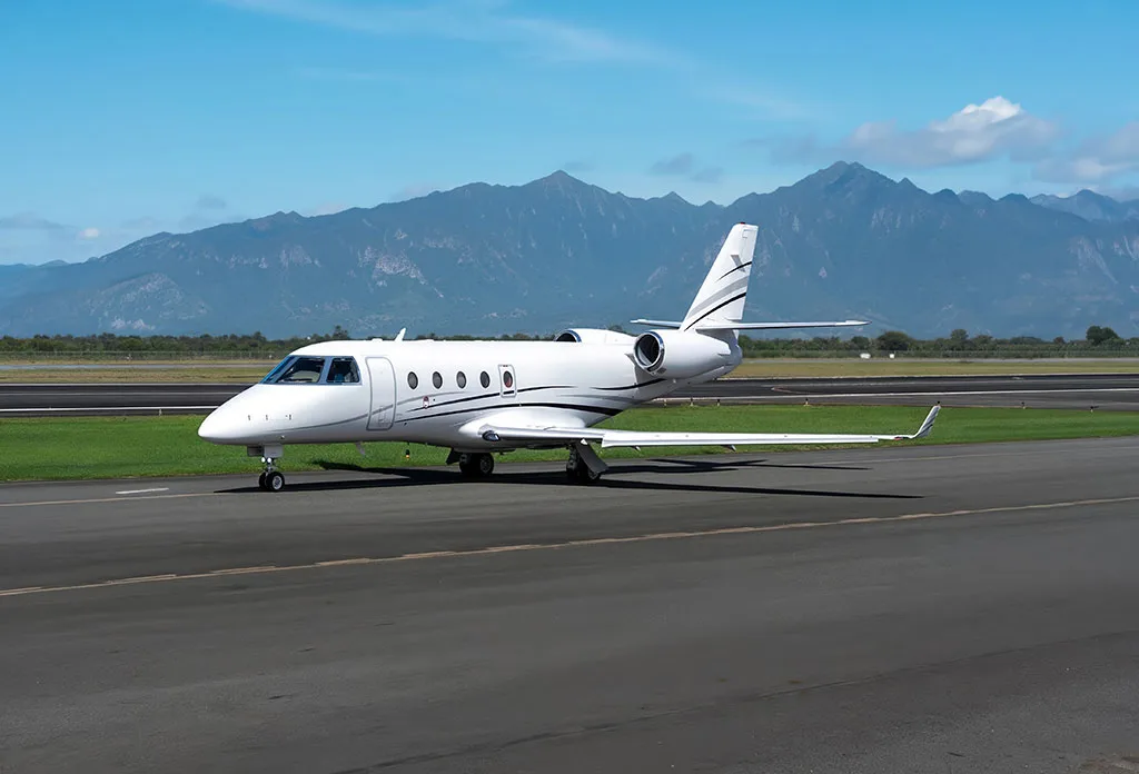 A white business jet with black and grey stripes is parked on a runway with mountains in the background.