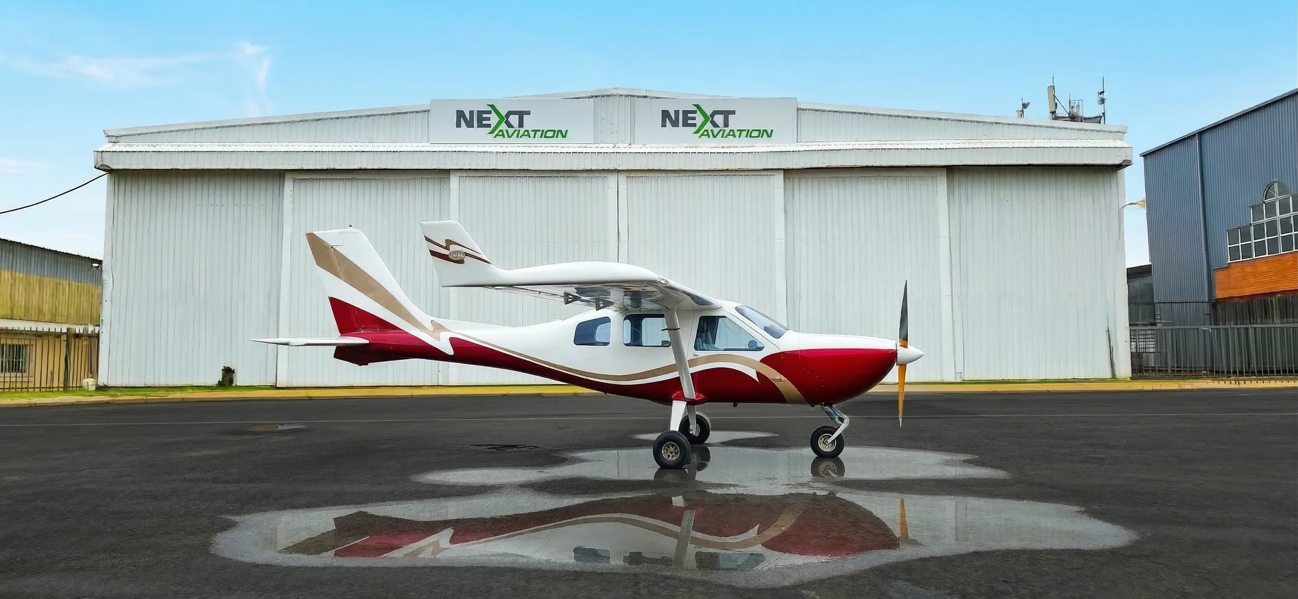 A white, red, and gold high-wing utility aircraft is parked on a wet tarmac in front of a hangar.
