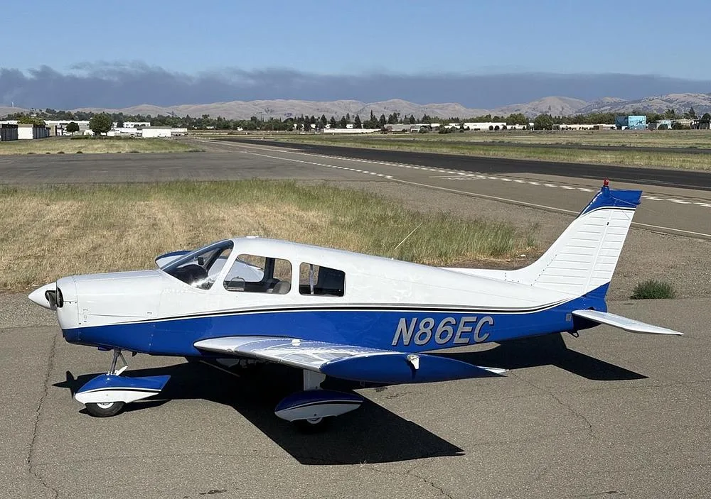 A white and blue Piper Cherokee aircraft (N86EC) parked on an airfield with a runway and distant hills.