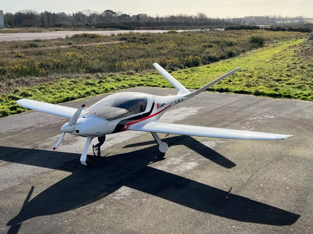A white Risen aircraft with red and black accents and registration F-JKLA parked on an asphalt runway.