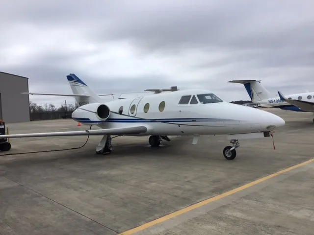 A white business jet with blue and grey stripes is parked on an airfield next to a hangar under a cloudy sky.