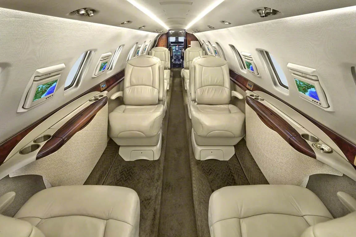 The spacious interior cabin of a private jet featuring beige leather seats, wood accents, and individual entertainment screens.