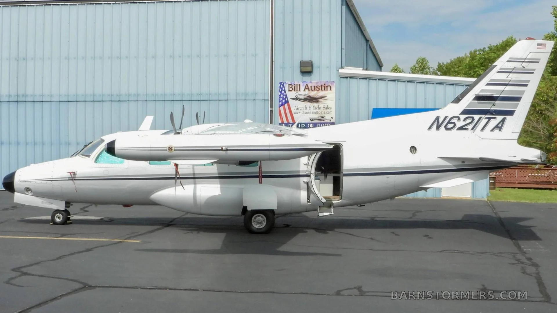 A white twin-engine turboprop aircraft, N621TA, with its passenger door open, parked on an asphalt apron next to a blue hangar.