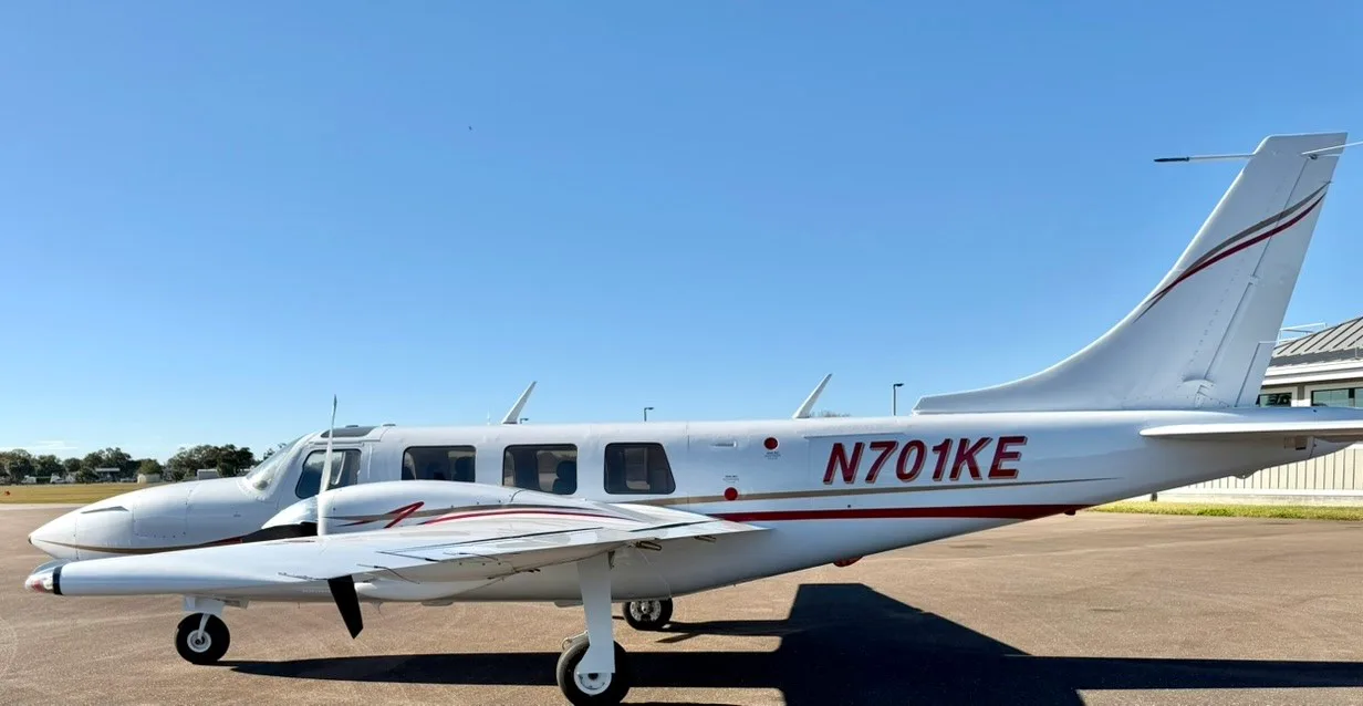 A white twin-engine propeller aircraft with red and gold stripes, registration N701KE, parked on an asphalt ramp under a clear blue sky.
