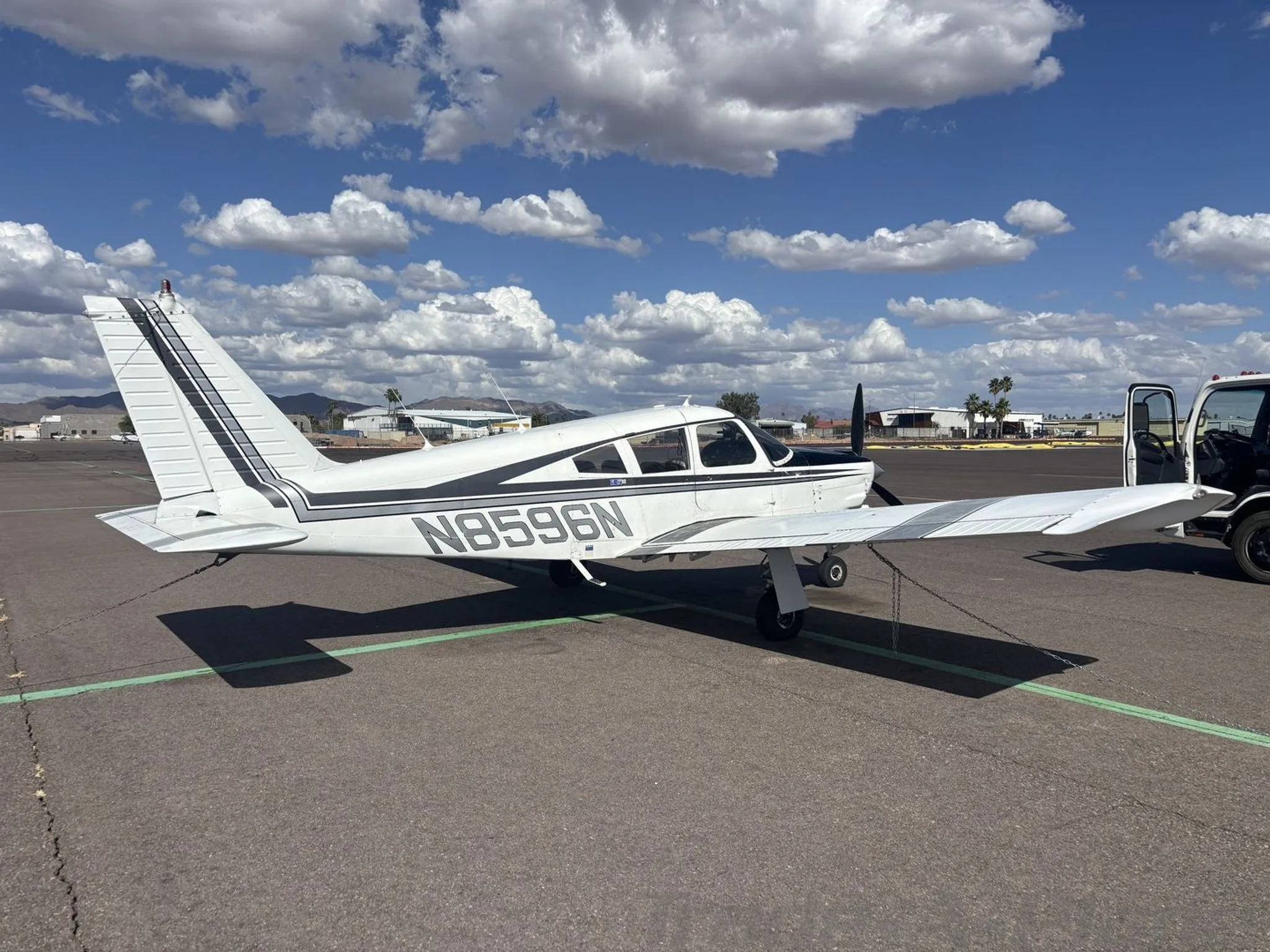 A white and black Piper Cherokee aircraft (N8596N) is parked on an airport tarmac next to a service vehicle.