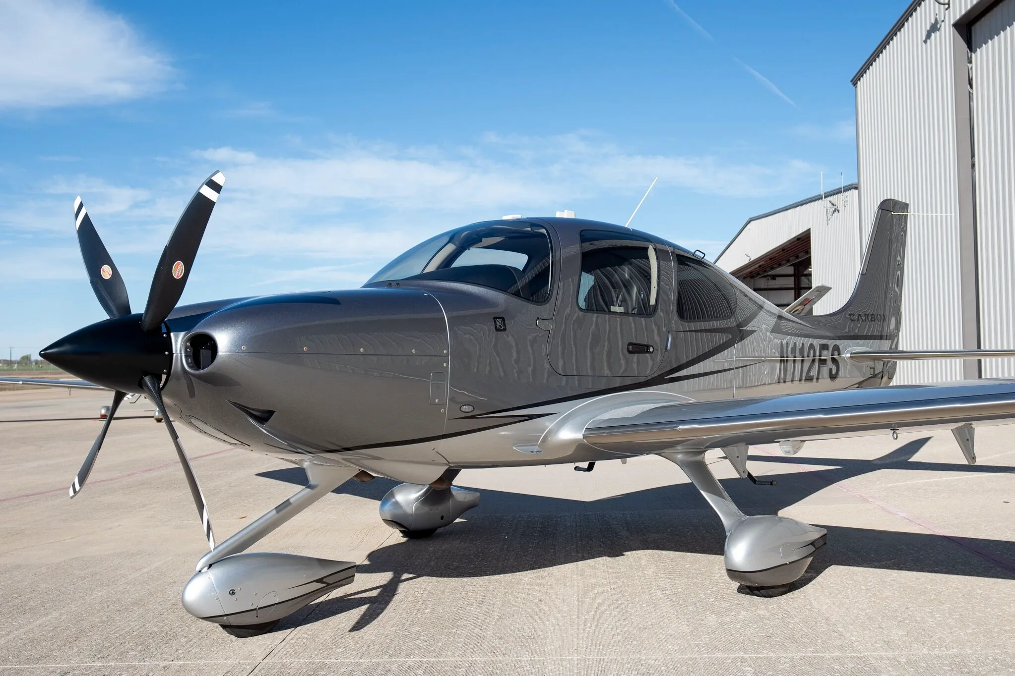 A front-left view of a silver and dark grey Cirrus SR22T Carbon Edition aircraft (N112FS) on an airport tarmac.