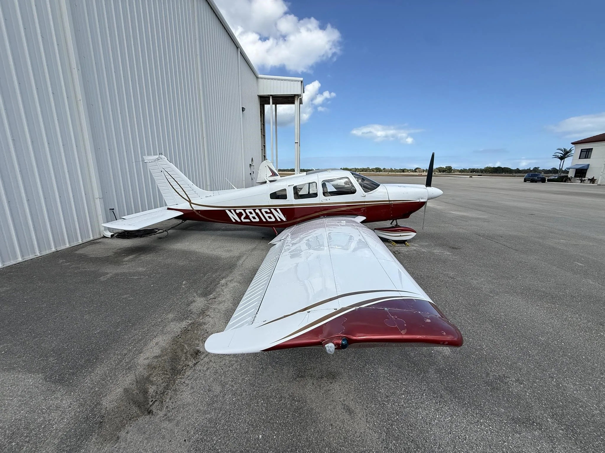 A white and maroon single-engine propeller aircraft, tail number N2816N, is parked on an asphalt apron next to a white hangar under a blue sky.