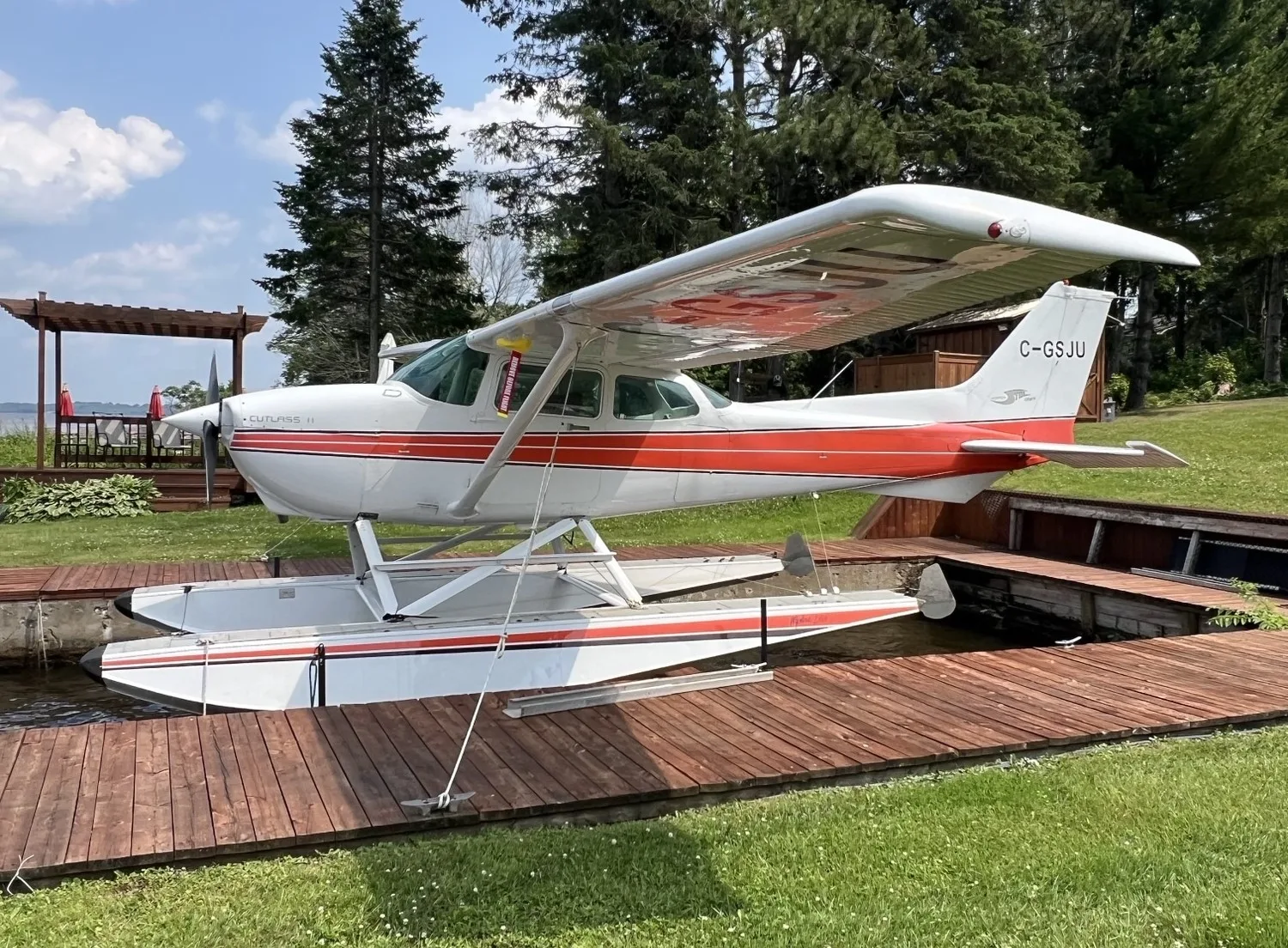 A white and red Cessna Cutlass II floatplane (C-GSJU) is parked on a wooden dock next to a lake.