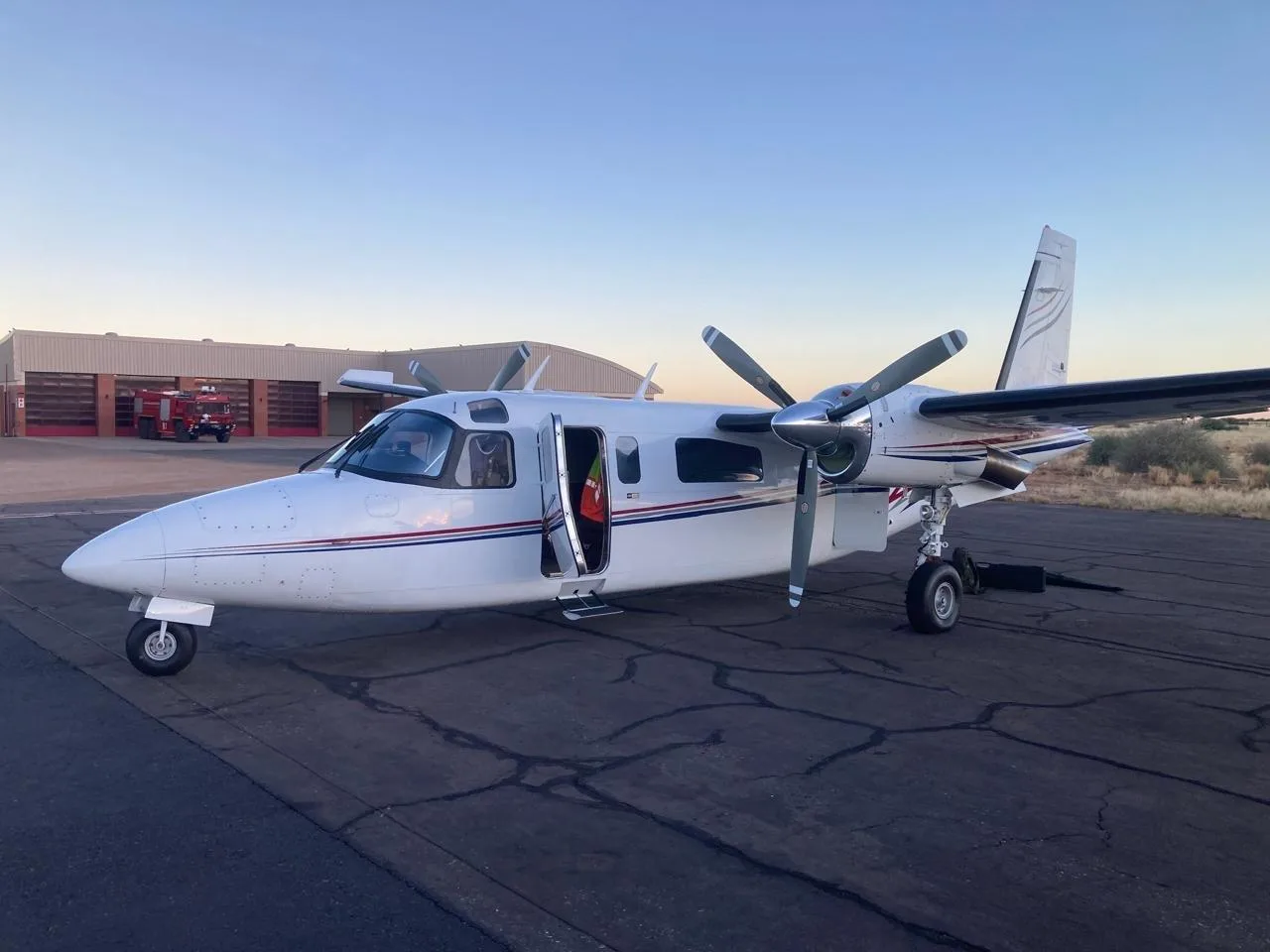 A white twin-engine turboprop aircraft with its passenger door open is parked on an airfield at dusk, with a red fire truck in the background.