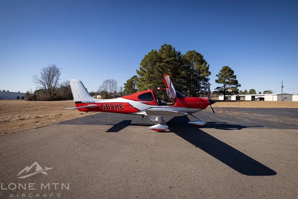 A red and silver Cirrus SR22T aircraft with open doors parked on an asphalt ramp with trees and buildings in the background.