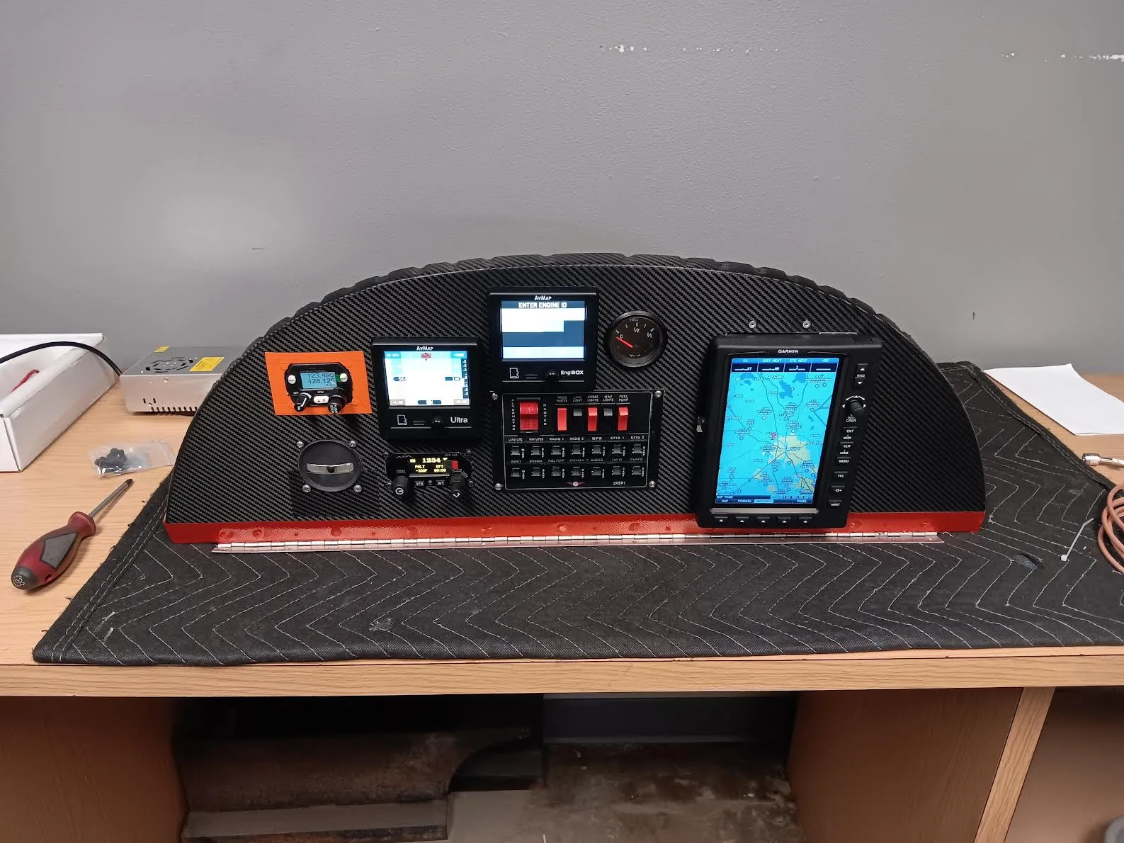 A standalone carbon fiber instrument panel with multiple digital displays and switches, resting on a workbench.