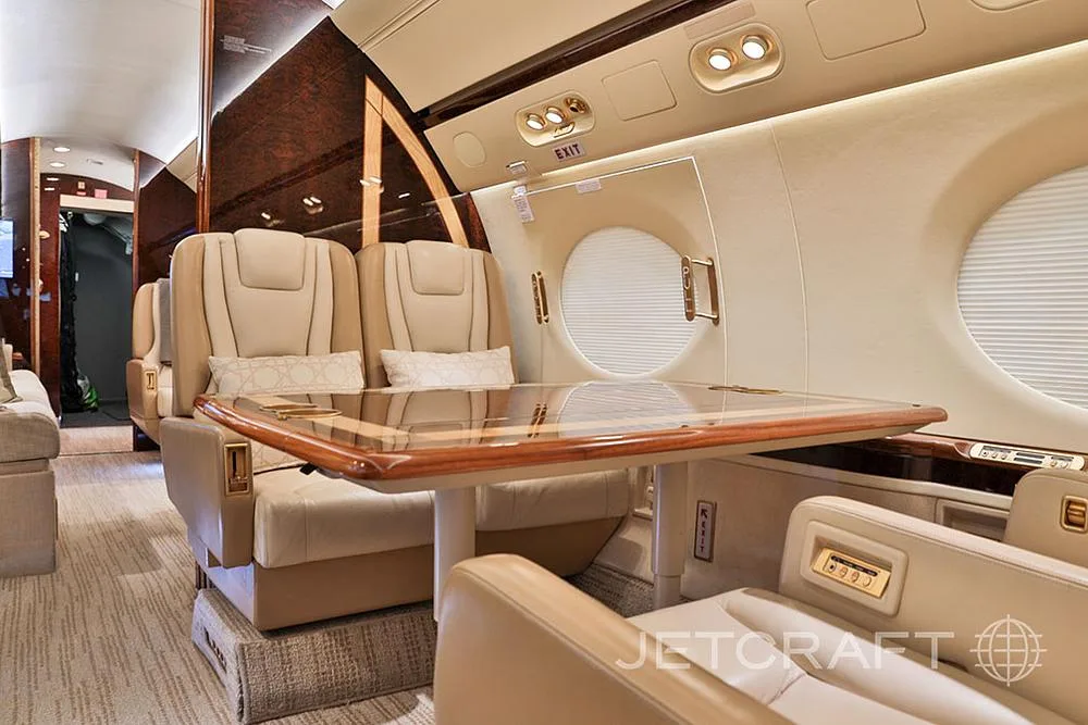 A close-up of the private jet's cabin, showing beige leather seats, a glass-top table, and a window with a closed shade.
