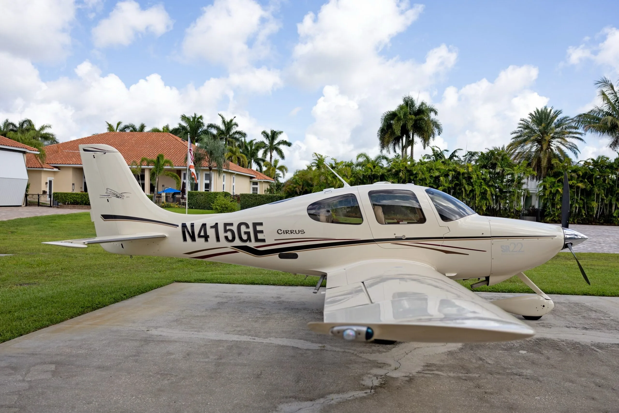 A white Cirrus SR22 aircraft with black and maroon stripes, registration N415GE, parked on a concrete pad with palm trees and houses in the background.