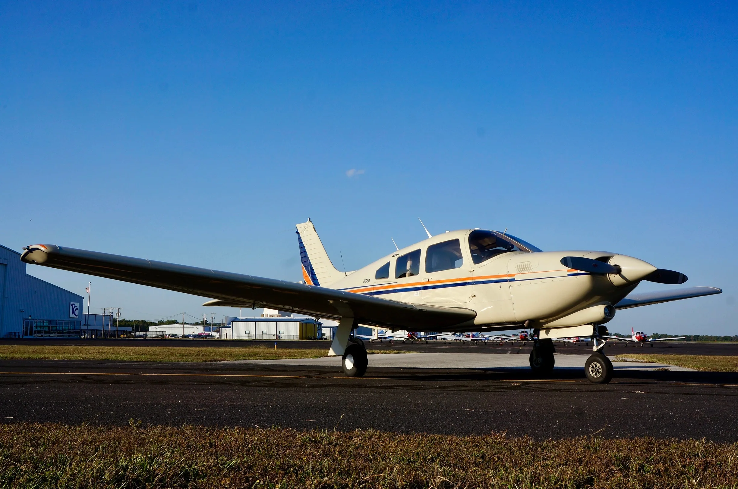 A cream-colored Piper aircraft with blue and orange stripes on an airport tarmac under a clear blue sky.