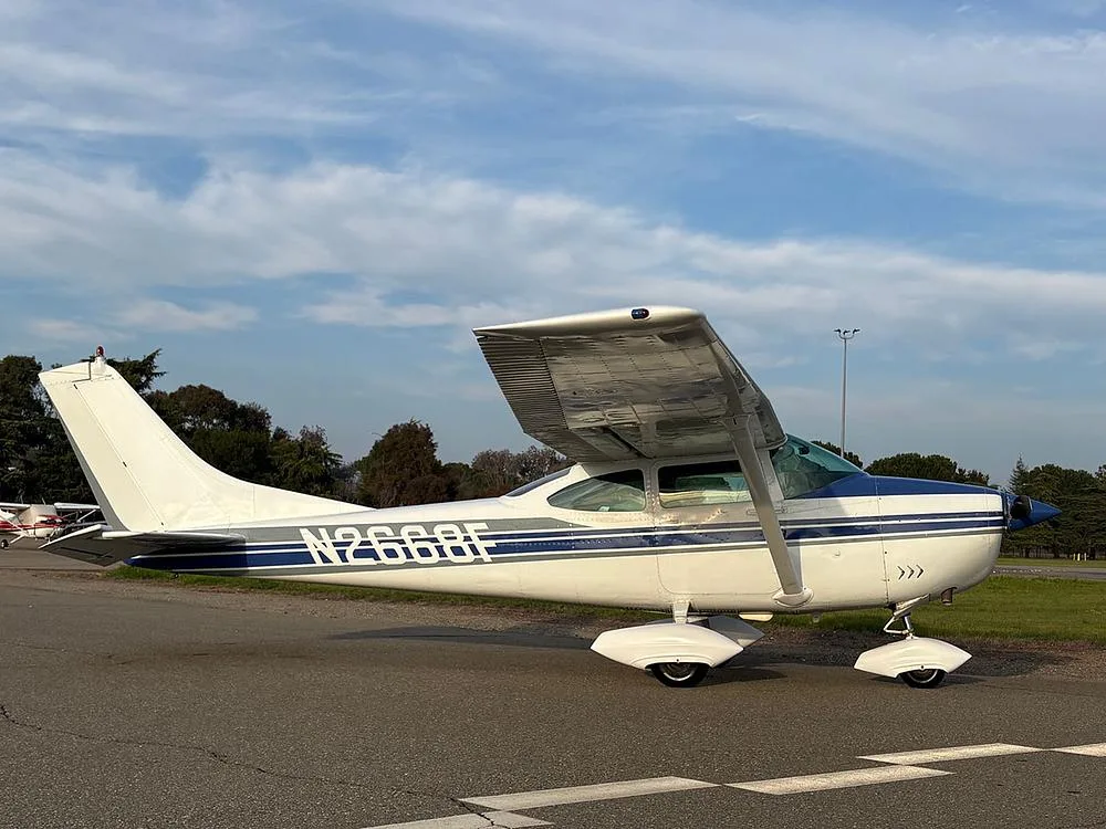 A white and blue Cessna 182 aircraft (N2668F) parked on an airfield under a partly cloudy sky.