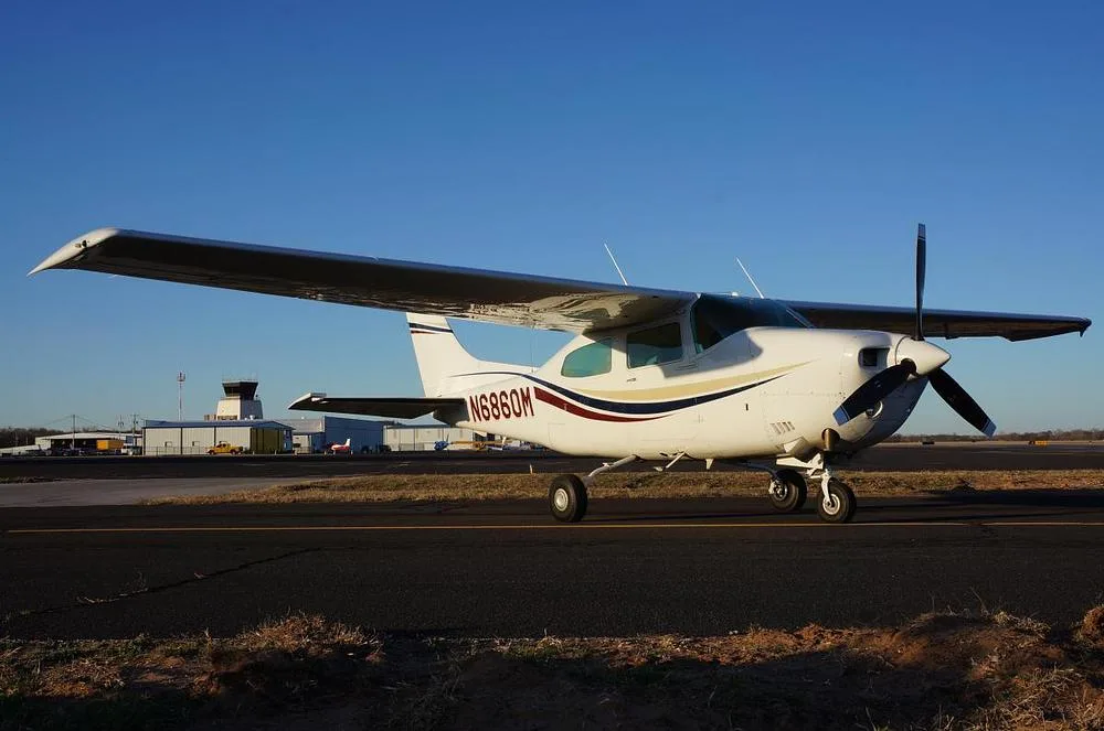 A white Cessna 210 Centurion aircraft with red, blue, and gold stripes on an airfield with a control tower in the background.