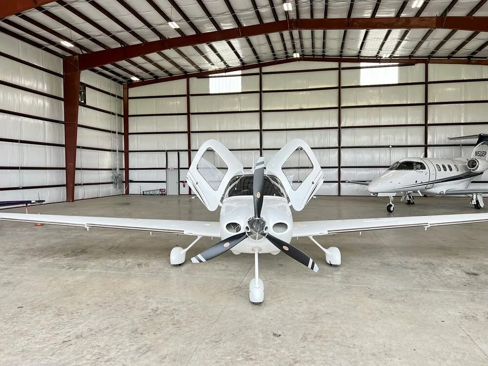 A white Cirrus SR22 aircraft with its doors open, parked in a hangar next to another jet.