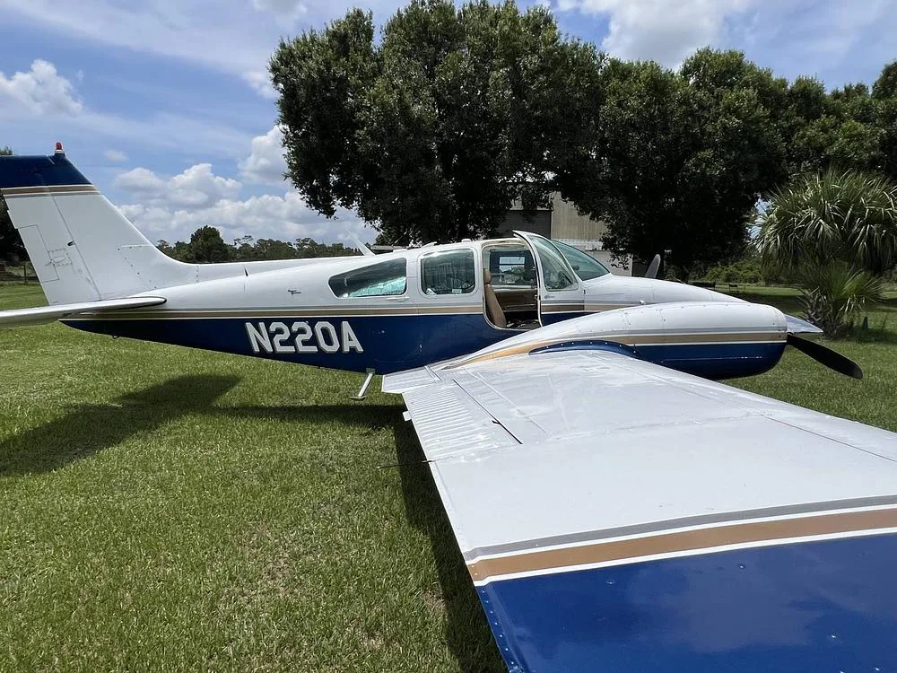 A white and blue twin-engine aircraft, N220A, with its door open, is parked on a grassy field.