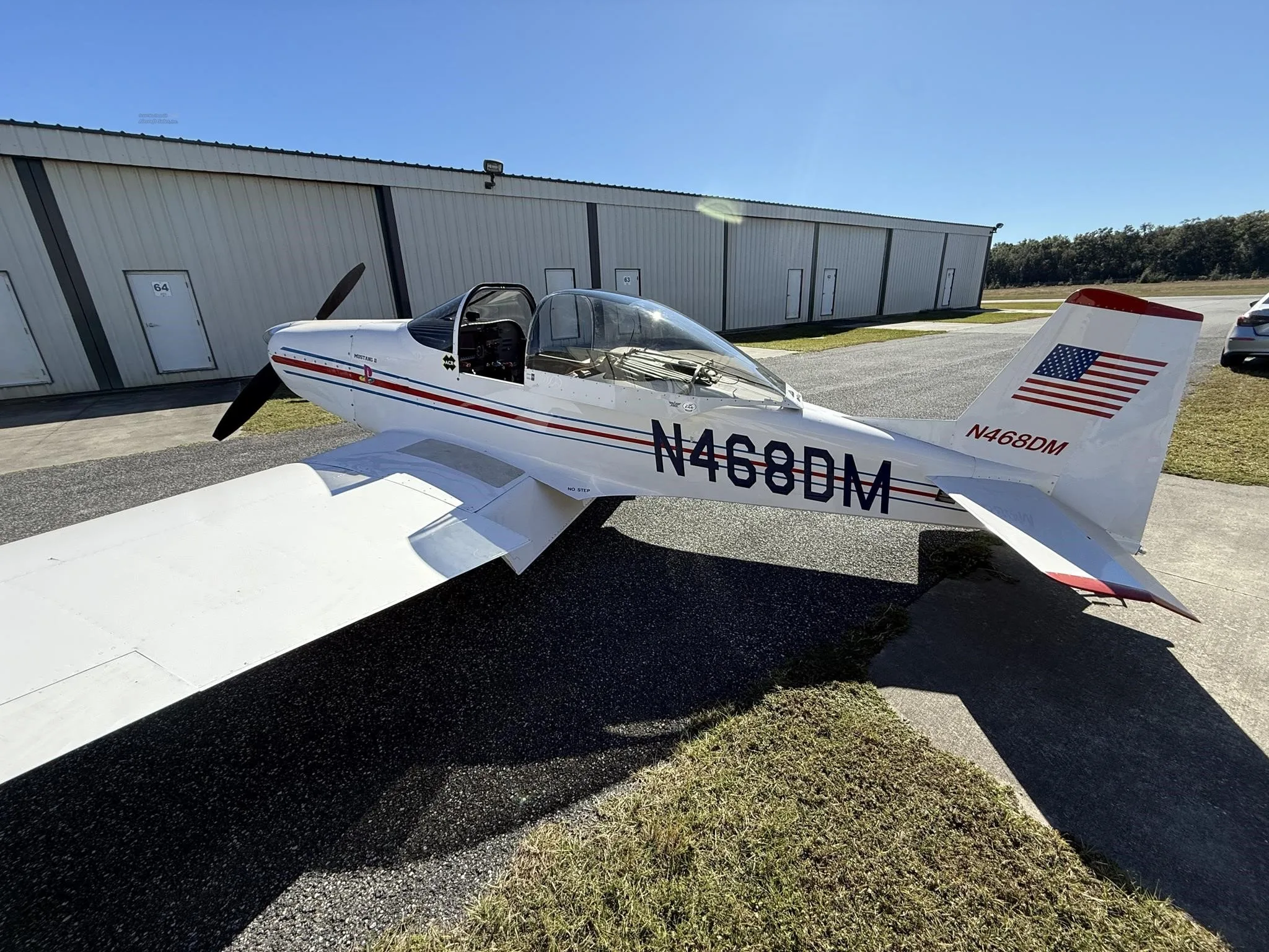 A white Mustang II aircraft with red and blue stripes, registration N468DM, parked on a tarmac next to hangars.