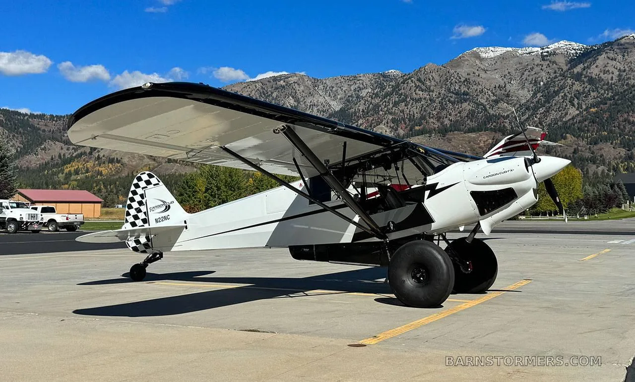 A white Carbon Cub FX-3 aircraft with black accents and large tundra tires is parked on an asphalt ramp with mountains in the background.