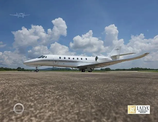 A white Cessna Citation business jet is parked on an asphalt runway under a partly cloudy blue sky, with another aircraft faintly visible in the distance.