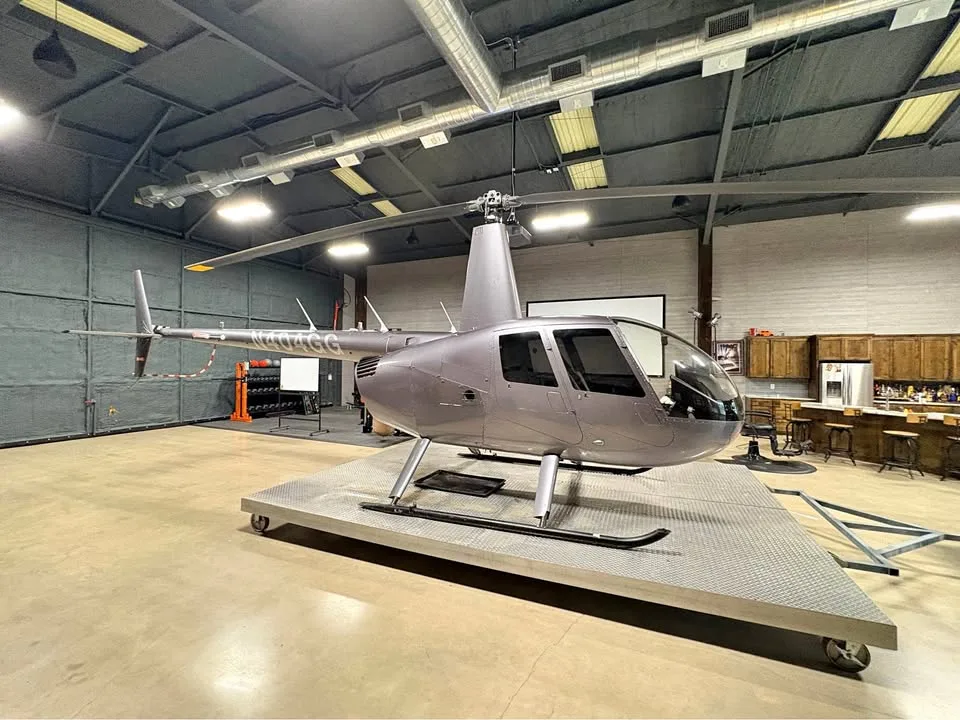 A grey Robinson R44 helicopter on a display platform inside a spacious hangar-like room.