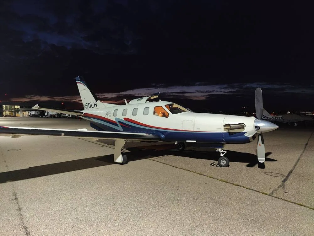 A TBM 850 aircraft with white, blue, and red livery parked on an airfield at dusk or night, with hangar lights in the background.