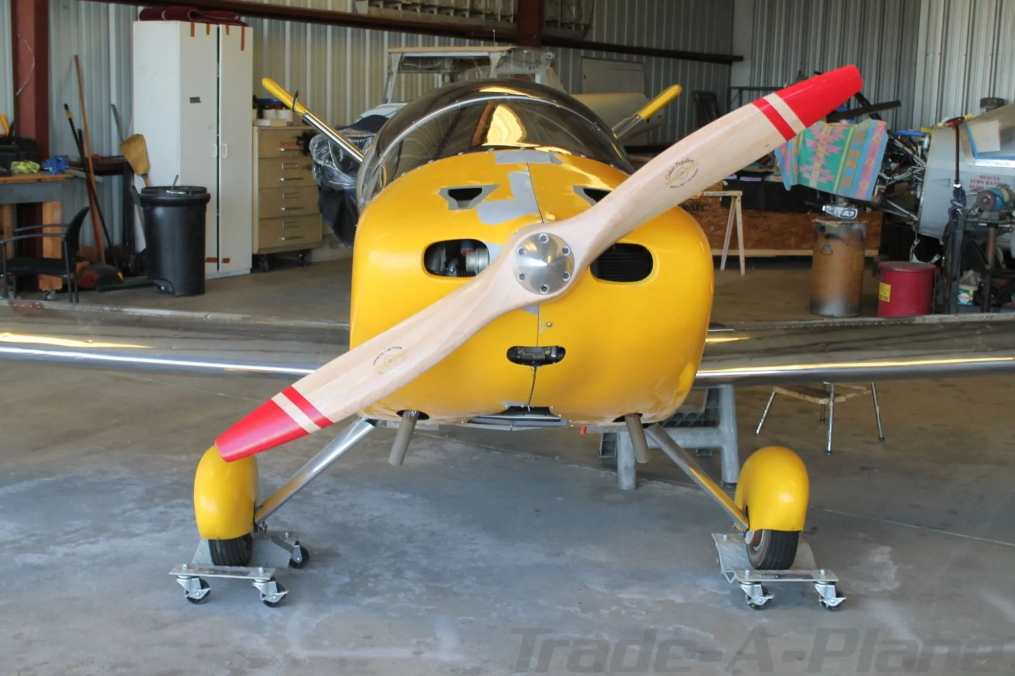 A partially assembled, unpainted metal aircraft with a yellow nose, wooden propeller, and yellow landing gear, viewed from the front in a hangar.