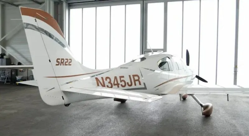 A rear-quarter view of a white Cirrus SR22 aircraft with brown and silver accents, registration N345JR, in a hangar.