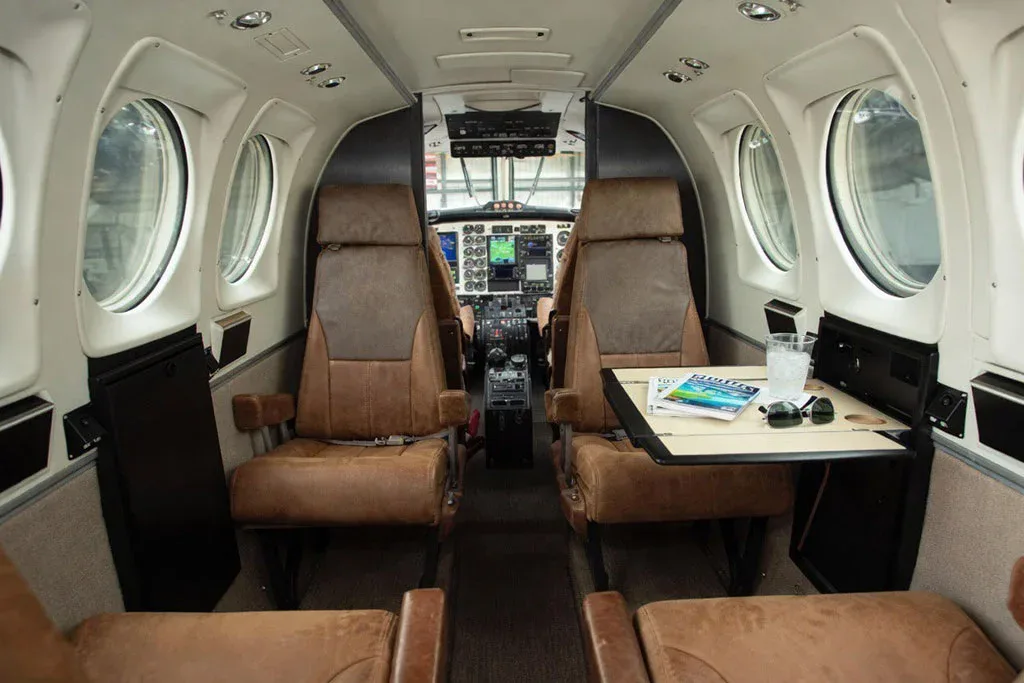 The interior cabin of a Beechcraft King Air, showing brown leather seats, a table with a drink, and the cockpit visible in the background.