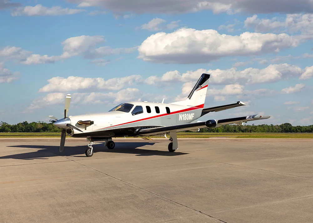 A white and grey TBM 900 series aircraft with red stripes, registration N180MF, parked on an airfield from a slightly different angle.