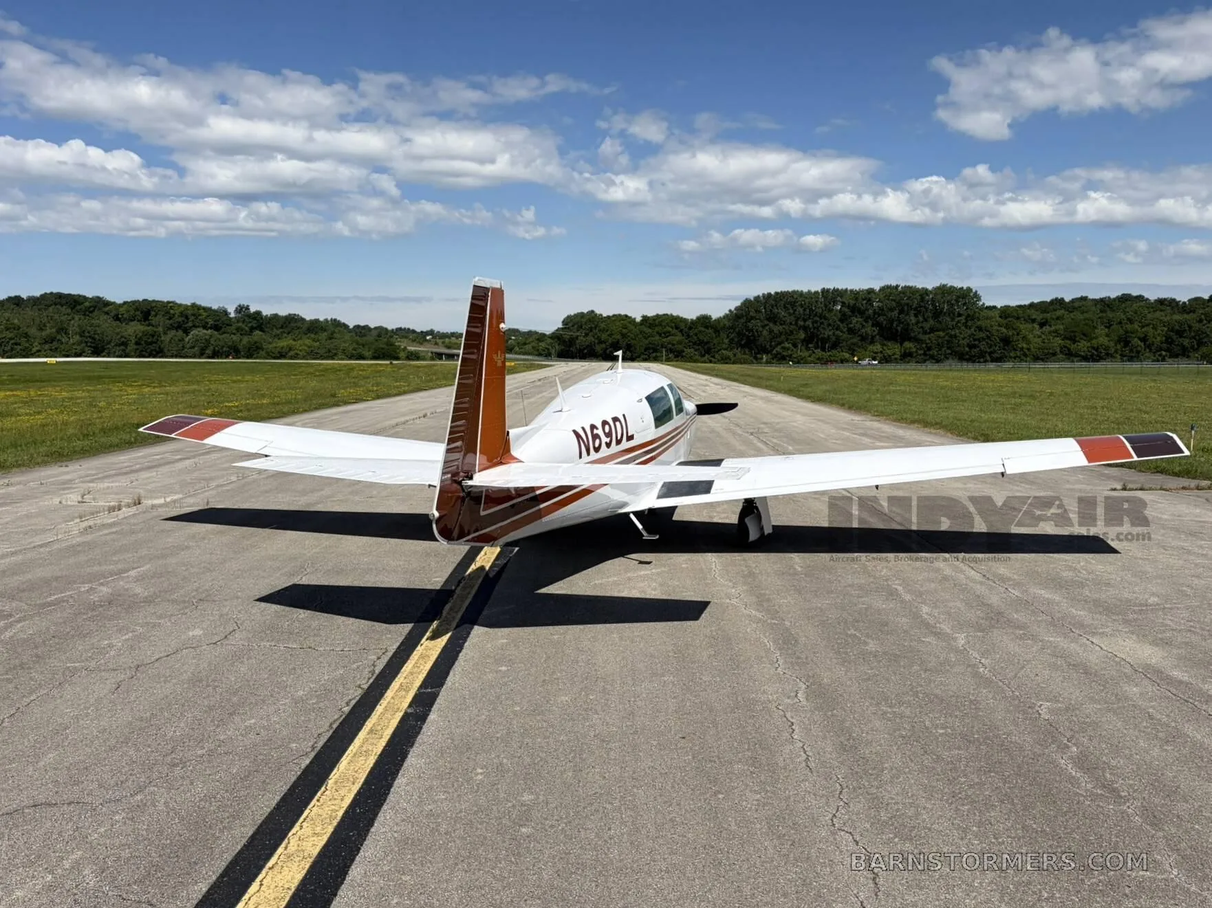 A white and brown Mooney M20J aircraft (N69DL) is parked on an asphalt runway under a partly cloudy sky, viewed from the rear.